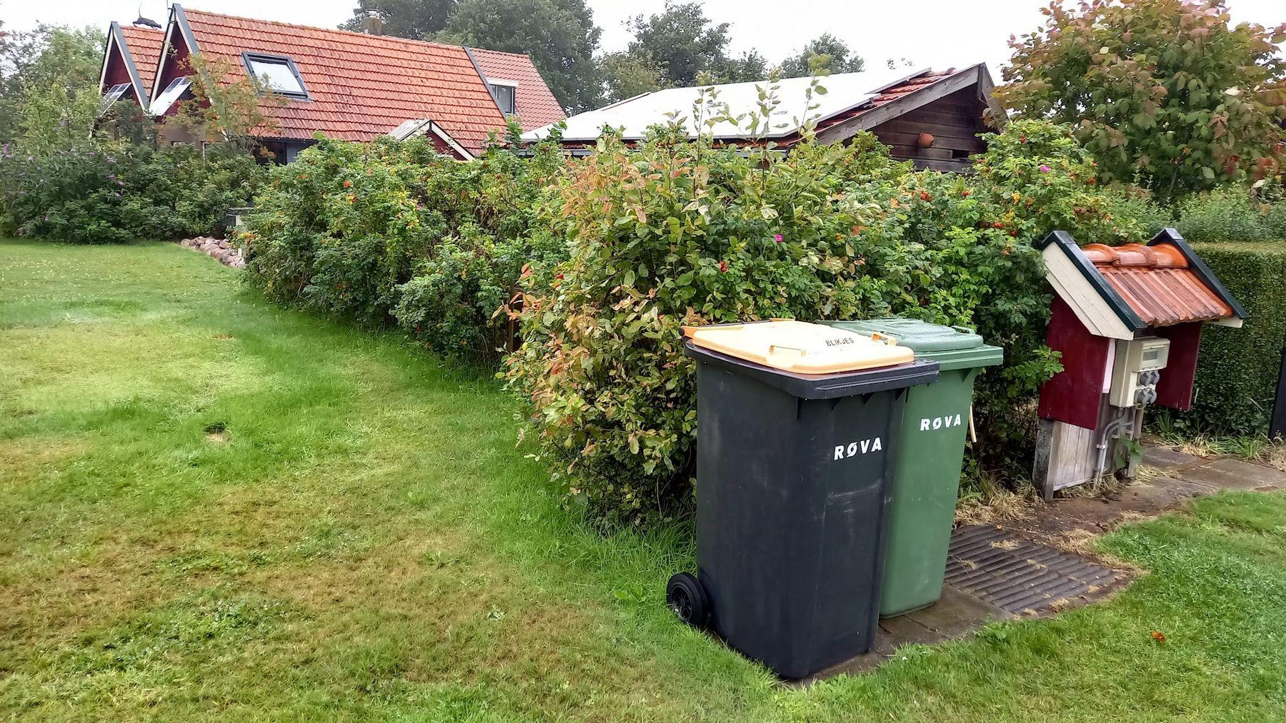 De rand van het veld met een rozenhaag, twee containers en een klein paaltje met een dakje, het huis erachter